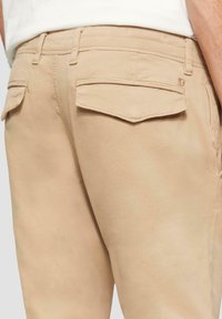Beige broek van een gestructureerde stof, met twee achterzakken en zichtbare stiksels. De tailleband heeft riemlussen.