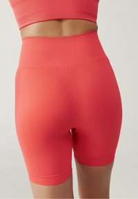 Vysoké korálové sportovní šortky vyrobené z texturované, elastické tkaniny. Mají přiléhavý střih, hladký pas a délku do poloviny stehna.