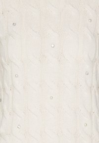 Bílá pletená tkanina s kabelovým vzorem zdobená malými perlami a jasnými skleněnými kamínky rozptýlenými po povrchu.