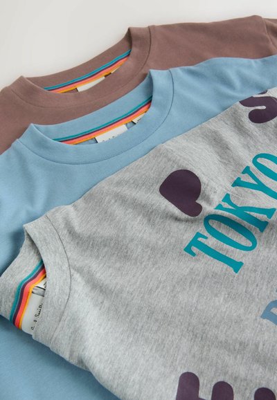 Trois T-shirts étalés à plat : marron, bleu clair et gris. Le T-shirt gris présente un texte graphique turquoise et des accents violet foncé. Tissu en coton.