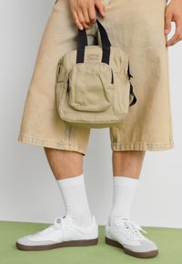 Inimene kannab beeže lühikesi pükse, valgeid sokke ja valgeid spordijalatsite, hoides väikest beeži seljakotti mustade käepidemetega enda kõrval.