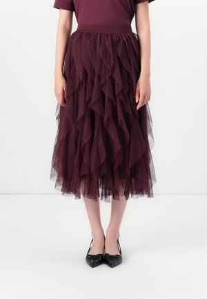 Kvinne iført en mørk lilla lagdelt tyll-midi skjørt og svarte spissede slingback hæler, stående mot en enkel hvit bakgrunn.