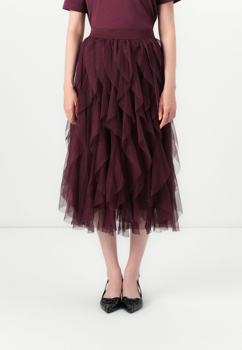 Donna che indossa una gonna midi in tulle a strati di un viola scuro e un paio di scarpe slingback nere con punta a punta, in piedi contro uno sfondo bianco semplice.