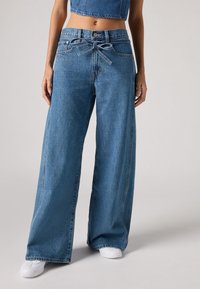 Calças de ganga de perna larga em um tom de azul claro, apresentando uma cintura atada e um design clássico de cinco bolsos com uma superfície ligeiramente texturizada.