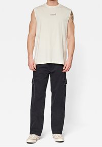 Maglietta senza maniche beige chiaro con piccolo logo nero "mavi", abbinata a pantaloni cargo neri con tasche laterali e vestibilità rilassata.