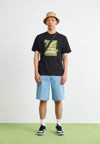 Black graphic t-shirt with yellow print, blue denim shorts, and black sneakers with white accents. Hat is beige and bucket-style.
