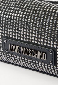 Black bag covered in silver circular studs, featuring a rectangular "LOVE MOSCHINO" logo patch. Smooth texture with shiny metallic accents.