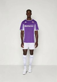 Violet futbola krekls ar baltām akcentiem, īsām piedurknēm, ar logotipu uz krūtīm, savienots ar baltām biksēm un ceļgala garuma zeķēm. Uz kājām sporta apavi.