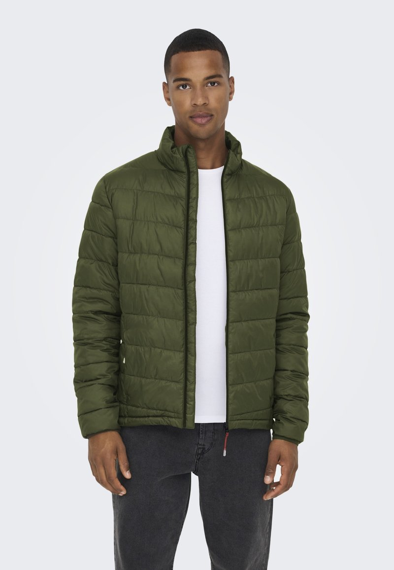 Groene pufferjack met een gestikt ontwerp, hoge kraag en een ritssluiting aan de voorkant. Gecombineerd met een wit overhemd, zwarte jeans en een slim fit.