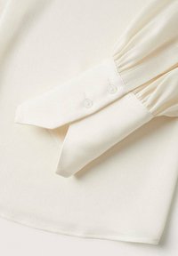 Cremfarbige Bluse aus strukturiertem Stoff mit langen, puffartigen Ärmeln und einem schmal zulaufenden Manschetten mit zwei Knopfverschlüssen.