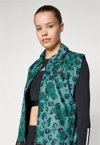 Veste sans manches à motif léopard vert avec un col, fermeture éclair et deux poches latérales, portée sur un haut noir. Tissu lisse et brillant.