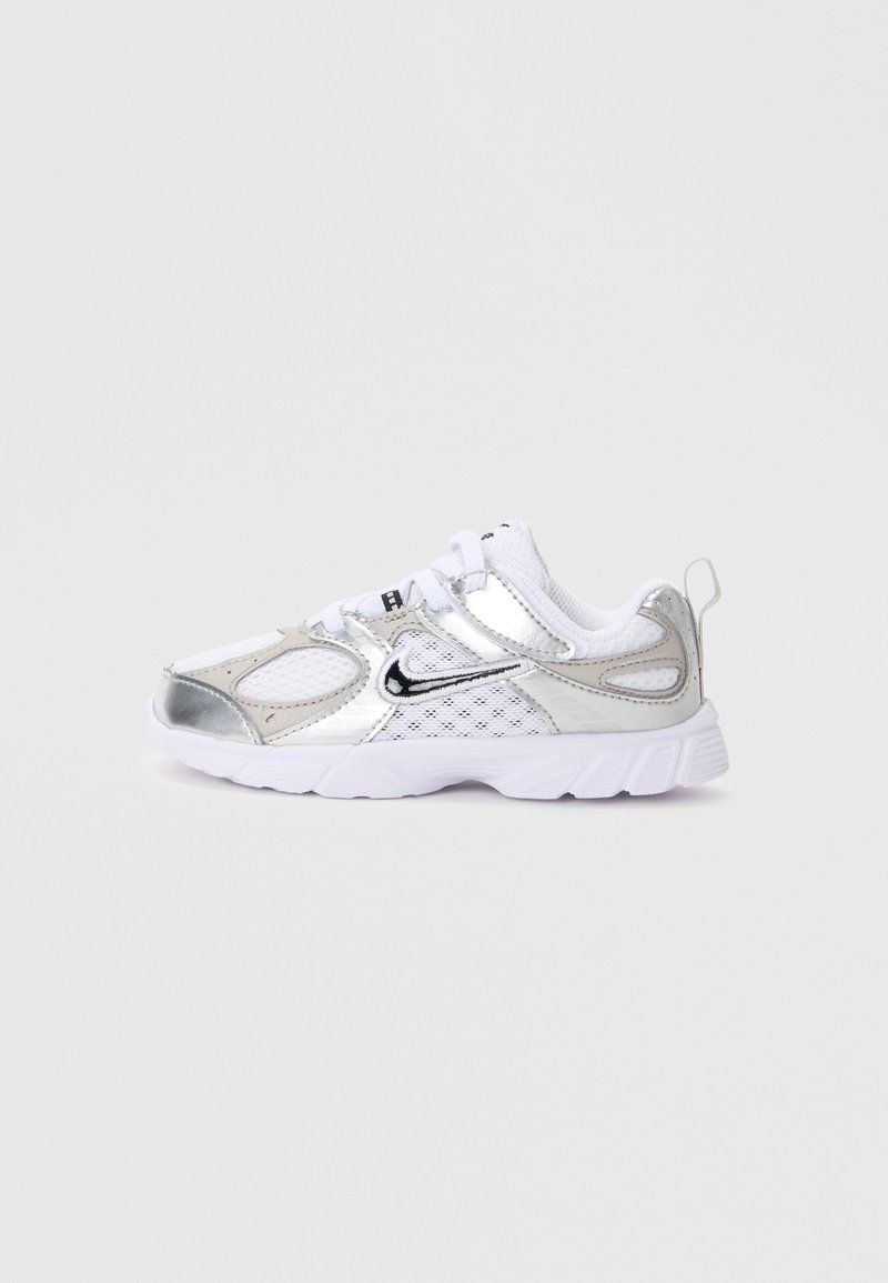 Baskets Nike blanches et argentées avec panneaux en mesh, logo swoosh noir, lacets blancs et semelle blanche épaisse sur un fond gris clair uni.