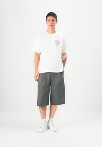 Hvit bomulls t-skjorte med et rødt grafisk motiv på venstre bryst, sammen med grå plissert shorts, iført hvite joggesko og grå sokker.