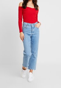Top rosso a spalle scoperte in maglia a coste, caratterizzato da maniche lunghe e un design aderente, abbinato a jeans azzurri a vita alta e sneakers bianche.