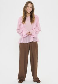 Helles rosa rüschengeschmücktes Blusenshirt mit langen Ärmeln und weit auslaufenden Manschetten, kombiniert mit lockeren braunen Hosen. Weicher, fließender Stoff mit geschichtetem Textur.