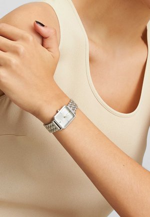 Montre en argent avec un cadran rectangulaire, bracelet en maillons métalliques et un design minimaliste. Portée sur un bras nu avec des vêtements aux teintes neutres.