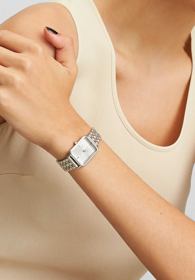 Orologio da polso argentato con quadrante rettangolare, bracciale a maglie metalliche e design minimalista. Indossato su un braccio nudo con abbigliamento dai toni neutri.