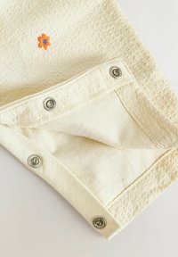 Tissu de couleur crème avec trois boutons-pression argentés le long du bord et une petite broderie de fleur orange près du haut.