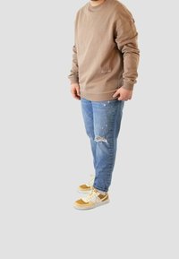 Hombre con sudadera marrón, vaqueros azules rasgados y zapatillas beige-blancas, de pie frente a un fondo liso.