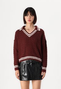 Maglione bordeaux a maglia con motivo a treccia, scollo a V e colletto a righe bianche; abbinato a una mini gonna nera in pelle con embossing a coccodrillo.