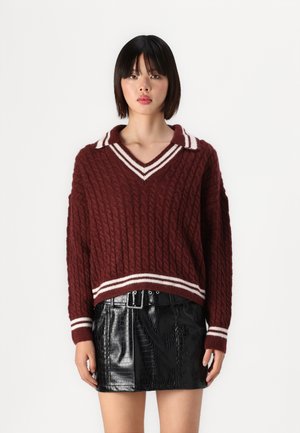 Maglione bordeaux a maglia con motivo a treccia, scollo a V e colletto a righe bianche; abbinato a una mini gonna nera in pelle con embossing a coccodrillo.