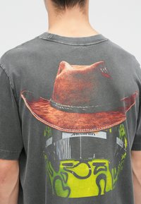 Graues T-Shirt mit einem großen Grafikdruck eines Cowboy-Huts über einem Rennhelm in Gelb und Grün, mit kunstvollen Details auf der Textur des Huts.