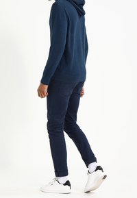 Felpa blu navy realizzata in tessuto morbido con tasca a marsupio; abbinata a pantaloni scuri a mezza gamba e sneakers bianche con accenti neri.