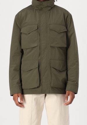Man die een olijfgroene jas met vier klepzakken draagt, staat tegen een effen achtergrond, gecombineerd met lichtbeige broek.