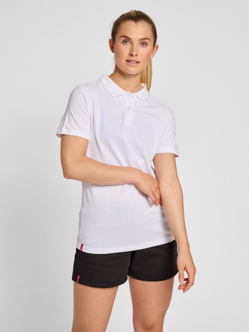 Vrouw met blond paardenstaart die een wit polohemd met korte mouwen en zwarte shorts draagt, en het shirtmouwtje aanpast tegen een effen grijze achtergrond.