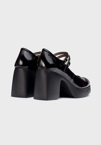 Zapatos de tacón de charol negro con puntera redondeada, dos correas ajustables y tacones gruesos, con un acabado liso y brillante.