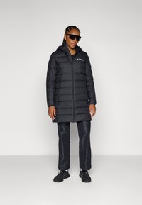 Black puffer coat with a zip closure and hood, featuring a logo on the chest. Worn with dark patterned pants and black shoes.