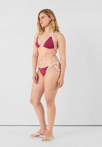 Set di bikini con reggiseno triangolare di un rosso intenso e slip coordinati con bordi bianchi e lacci laterali; il modello indossa ciabatte bianche.