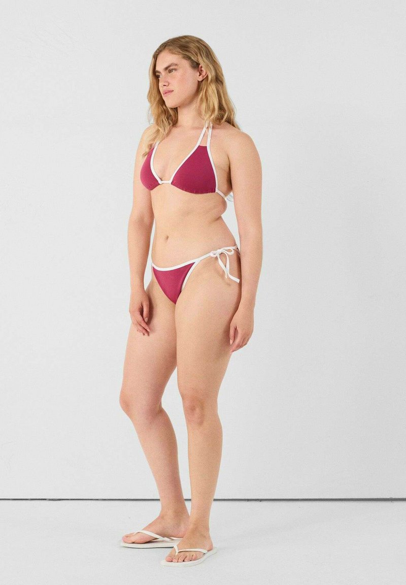 Set di bikini con reggiseno triangolare di un rosso intenso e slip coordinati con bordi bianchi e lacci laterali; il modello indossa ciabatte bianche.