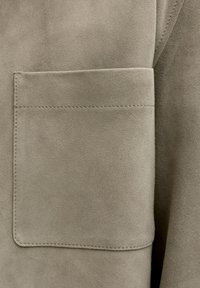 Κοντινή λήψη μπεζ σουέντ υφάσματος με ραμμένη ορθογώνια τσέπη στην αριστερή πλευρά.