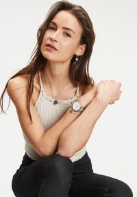 Silberne Uhr mit rundem Zifferblatt, Mesh-Armband; geschichtete Halskette mit Anhänger; gemusterte Ohrringe; Model trägt einen gestrickten beigen Pullover und schwarze Hose.