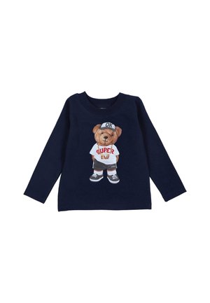 Marineblaues Langarmshirt mit einem Bärenmotiv auf einem weißen Shirt mit dem Text "SUPER BOY", graue Shorts und Sneakers sowie einer Mütze mit der Aufschrift "OK".