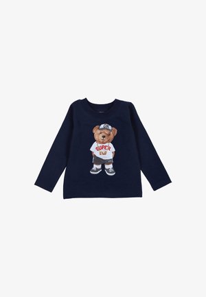 Marineblaues Langarmshirt mit einem Bärenmotiv auf einem weißen Shirt mit dem Text "SUPER BOY", graue Shorts und Sneakers sowie einer Mütze mit der Aufschrift "OK".