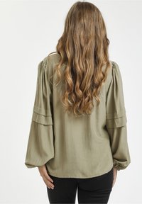 Olivgrüne Bluse mit langen, gepufften Ärmeln und gestuften Rüschen an den Schultern. Hergestellt aus einem leichten Stoff mit glatter Textur.
