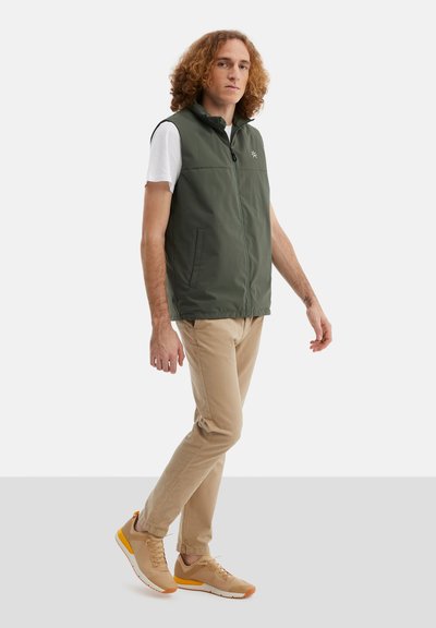 Tmavě zelená, bezrukávová vesta s vysokým límcem, černým zipem a bočními kapsami, nošená přes bílou košili. V kombinaci s béžovými kalhotami a teniskami.