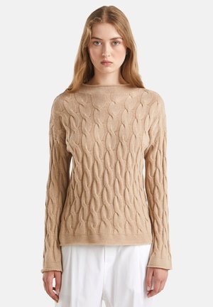 Jeune femme aux cheveux longs portant un pull en tricot torsadé beige et un pantalon blanc, se tenant devant un fond uni.