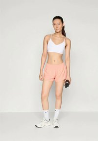 Soutien-gorge de sport blanc, shorts roses avec logo noir, et baskets blanches. Le mannequin tient une bouteille d'eau noire. Design simple et athlétique.