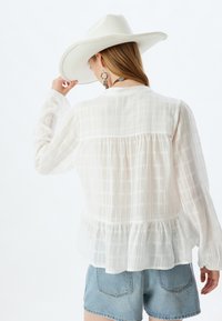 Weiße Bluse mit langen Ärmeln, strukturiertem, kariertem Muster, gerafftem Saum und Knöpfen an der Rückseite. Kombiniert mit Denim-Shorts.