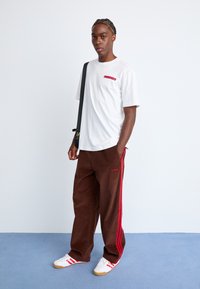 Hvid bomulds T-shirt med rødt logo, parret med brune corduroypants med røde sidelændere, komplet med hvide sneakers med røde accenter.