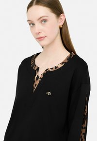 Giovane donna con capelli castani lisci indossa un top nero con accenti in stampa leopardata sul colletto e sulle maniche, e orecchini a forma di cuore dorati.