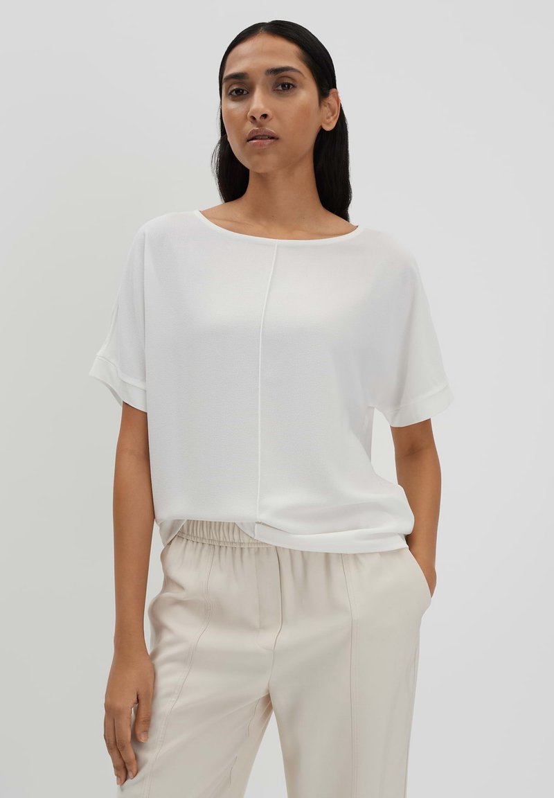 Blouse blanche à manches courtes avec une coupe ample, dotée d'un col rond et d'une couture subtile au centre. Associée à un pantalon beige clair.