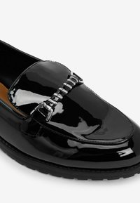 Zwarte gepatineerde leren loafers met een platte hak, voorzien van een decoratieve zilveren ketting aan de voorkant en een glanzende, gladde afwerking.