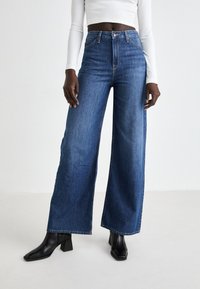 Donna con jeans blu a gamba larga a vita alta, top corto bianco a maniche lunghe e stivali neri con tacco quadrato in piedi su uno sfondo neutro.