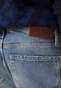 Hellblaue Denim-Jeans mit einem Lederetikett, das das "Pepe Jeans"-Logo zeigt, sichtbaren Nähten und zwei Gesäßtaschen.