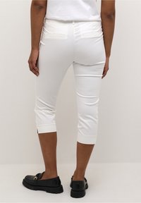 Pantalon blanc court et ajusté, avec poches arrière, ourlets retroussés et texture lisse. Porté avec des mocassins noirs.
