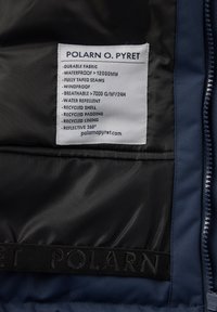 Mørkeblå jakke indvendigt med et label, der beskriver vandtæt stof, åndbare materialer, genanvendte materialer og reflekterende egenskaber. Tekstureret foring synlig.